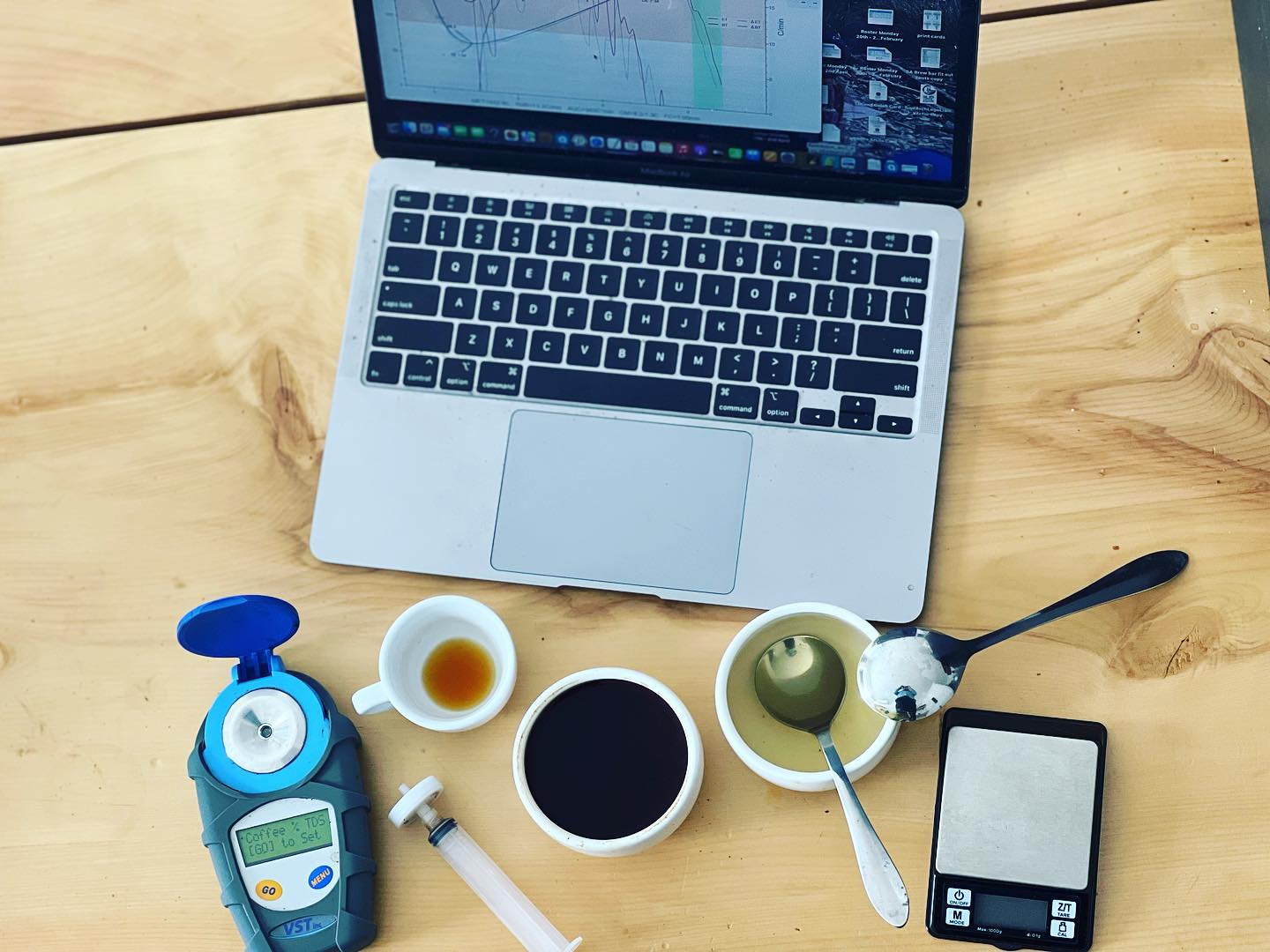 A wooden bench with a laptop showing roast curves, a TDS refractometer, cupping bowls, an espresso cup and a scale